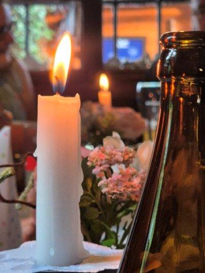 Eine brennende Kerze steht auf einem Tisch, umgeben von Blumen und einem Flaschenhals im Vordergrund.