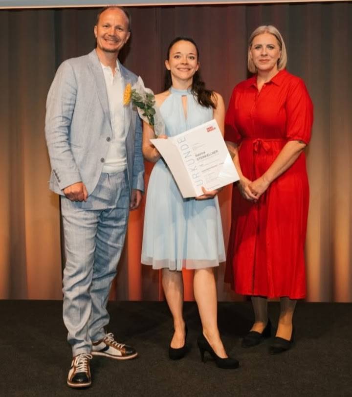 Eine Person erhält eine Auszeichnung während einer Zeremonie, umgeben von zwei anderen Personen.