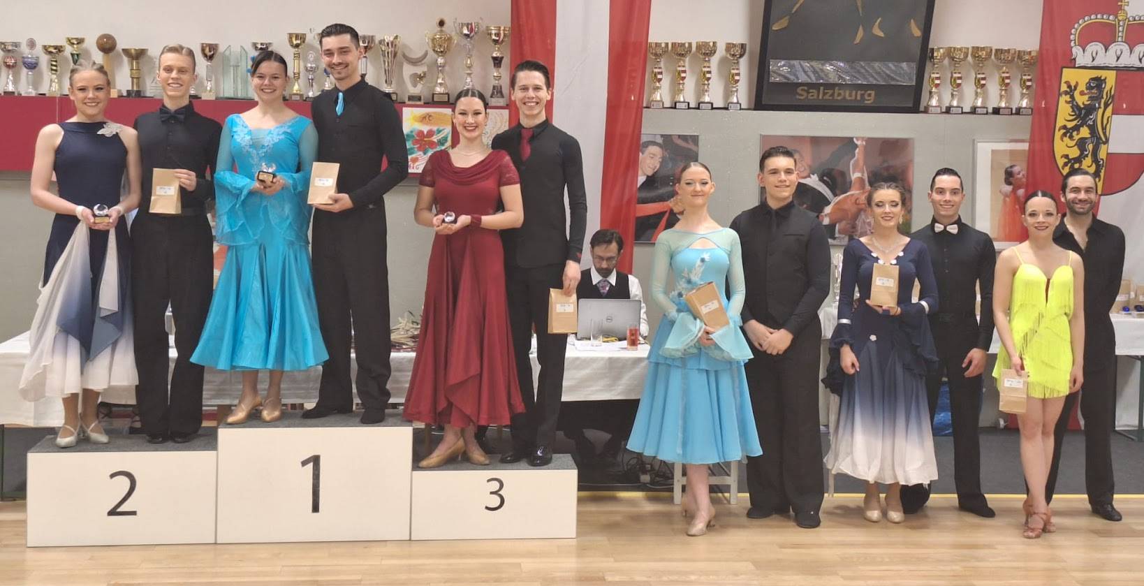 Eine Tanzwettbewerbssiegerehrung mit mehreren Paaren auf dem Podium und Pokalen im Hintergrund.