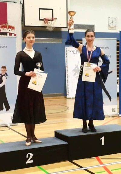 Zwei Tänzerinnen stehen auf einem Siegertreppchen, wobei die Gewinnerin mit einem Pokal und einer Urkunde in der Hand posiert.